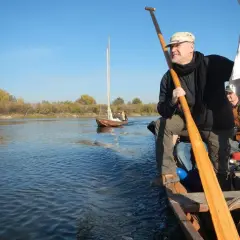 Mężczyzna z wiosłem na łodzi, obok płynie druga łódź. W tle jesienne drzewa i błękitne niebo. Wspaniała przygoda na dulki.