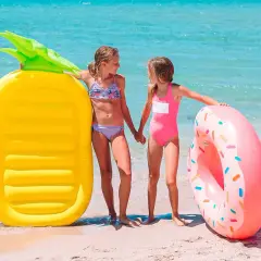 Dwie dziewczynki trzymają się za ręce na plaży, obok nich leży żółty materac do pływania w kształcie ananasa i różowa dmuchana oponka.