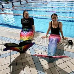 Dwie kobiety w kolorowych ogonach syreny, gotowe do mermaidingu, siedzą na brzegu basenu.