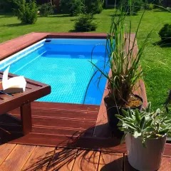 Relaks przy basenie stelażowym wkopanym w ziemię. Drewniany taras, leżak z książką i zielone rośliny tworzą oazę spokoju.