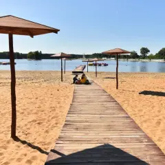 Sielpia jezioro w letni dzień. Drewniany pomost prowadzi na piaszczystą plażę, gdzie pod parasolami odpoczywają ludzie.