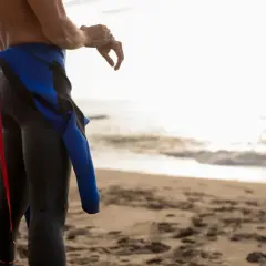 Mężczyzna w czarnej piance do pływania z niebieskimi wstawkami stoi na plaży, przygotowując się do wejścia do wody.