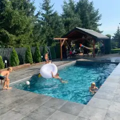 Dzieci bawią się w basenie w ziemi, jeden chłopiec próbuje zagrać w piłkę, inny pływa na białej dmuchanej poduszce.