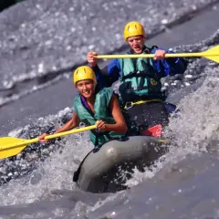 Dwóch uśmiechniętych ludzi w kaskach i kamizelkach płynie pontonem po rwącej rzece. To właśnie rafting – ekscytująca przygoda!