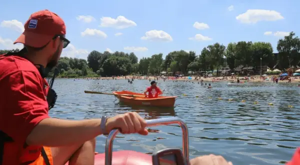 Ratownik wodny w łodzi obserwuje plażowiczów. W tle tłumy ludzi na plaży i w wodzie.