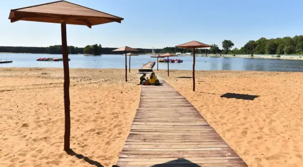 Sielpia jezioro w letni dzień. Drewniany pomost prowadzi na piaszczystą plażę, gdzie pod parasolami odpoczywają ludzie.