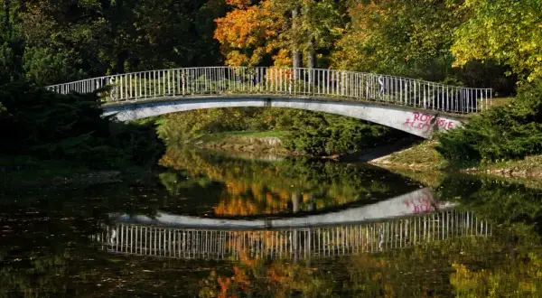 Mostek nad spokojną rzeką, odbijający jesienne barwy drzew. W oddali widać ludzi spacerujących po moście.