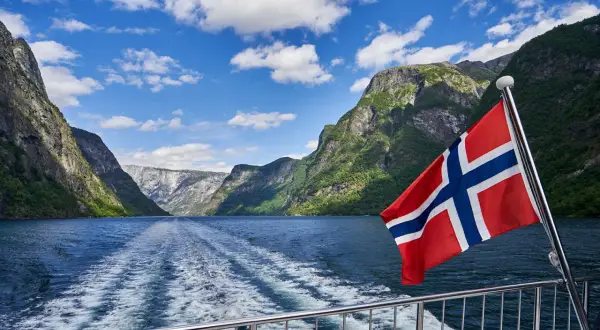 Norweska bandera flaga powiewa na tle malowniczego fiordu. Statek pruje wodę, zostawiając za sobą pianę.