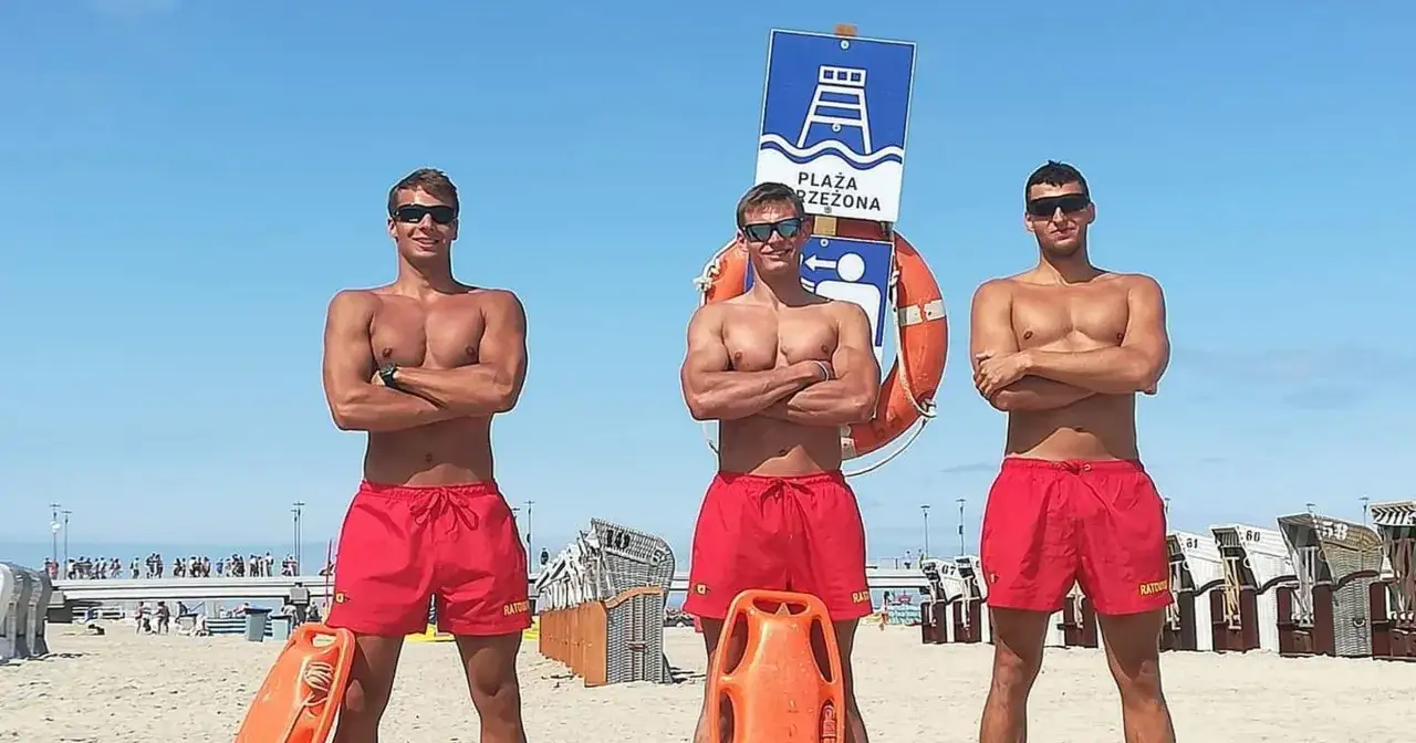 Trzech muskularnych ratowników wodnych w czerwonych kąpielówkach stoi na plaży.