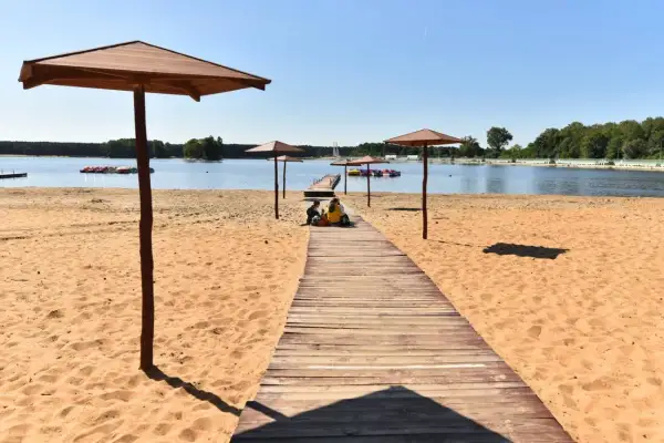 Sielpia jezioro w letni dzień. Drewniany pomost prowadzi na piaszczystą plażę, gdzie pod parasolami odpoczywają ludzie.