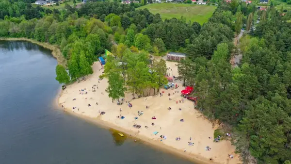 Plaża nad zalewem Cedzyna tętni życiem. Ludzie odpoczywają na piasku, ciesząc się słońcem i widokiem na jezioro.