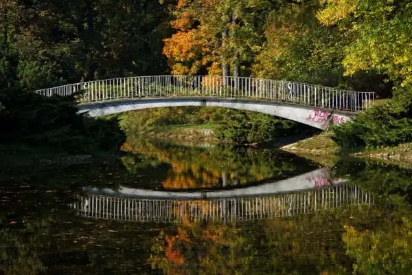 Mostek nad spokojną rzeką, odbijający jesienne barwy drzew. W oddali widać ludzi spacerujących po moście.